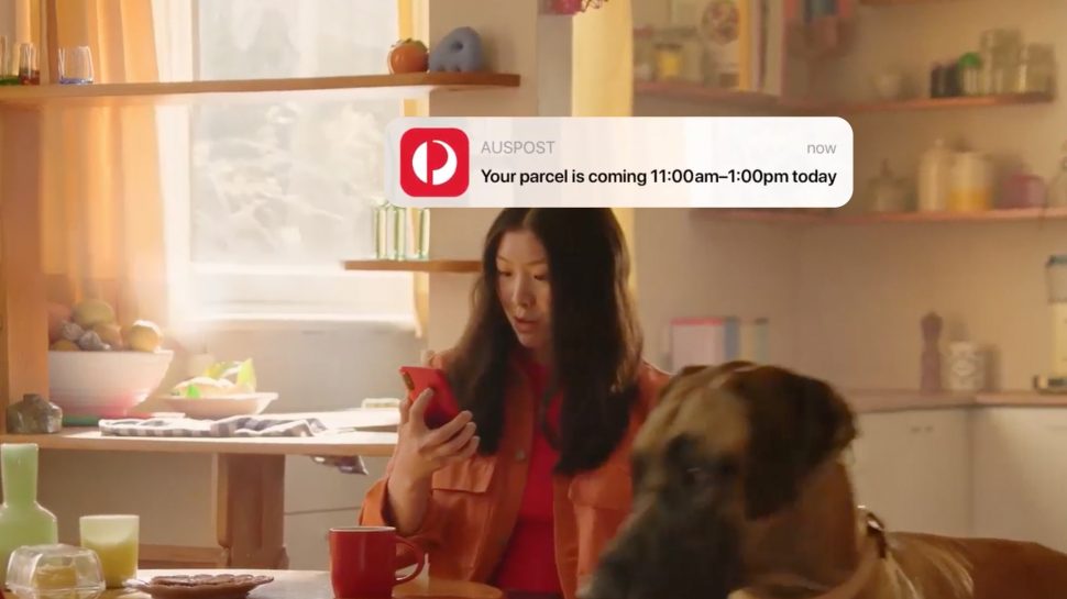 A woman checks her phone at the kitchen table. A notification from the Australia Post app shows the message "your parcel is coming 11:00am - 1:00pm today".